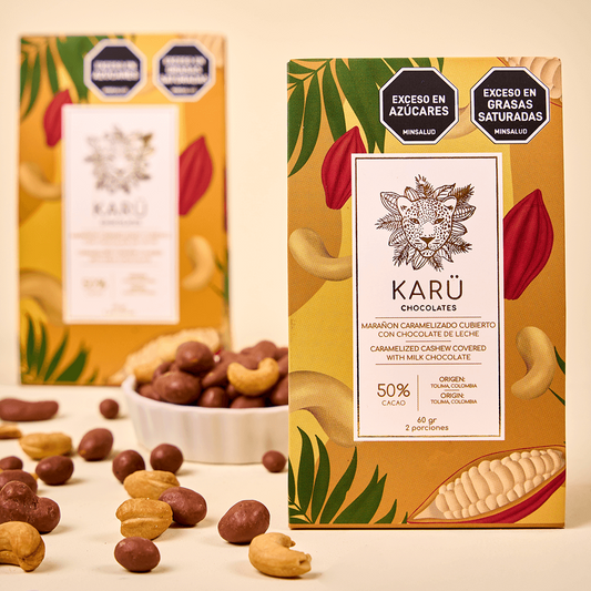 Two Karü chocolate-covered cashew boxes, one behind the other, with a bowl of chocolate-covered cashews in the center. Some cashews and chocolates are scattered on the surface.